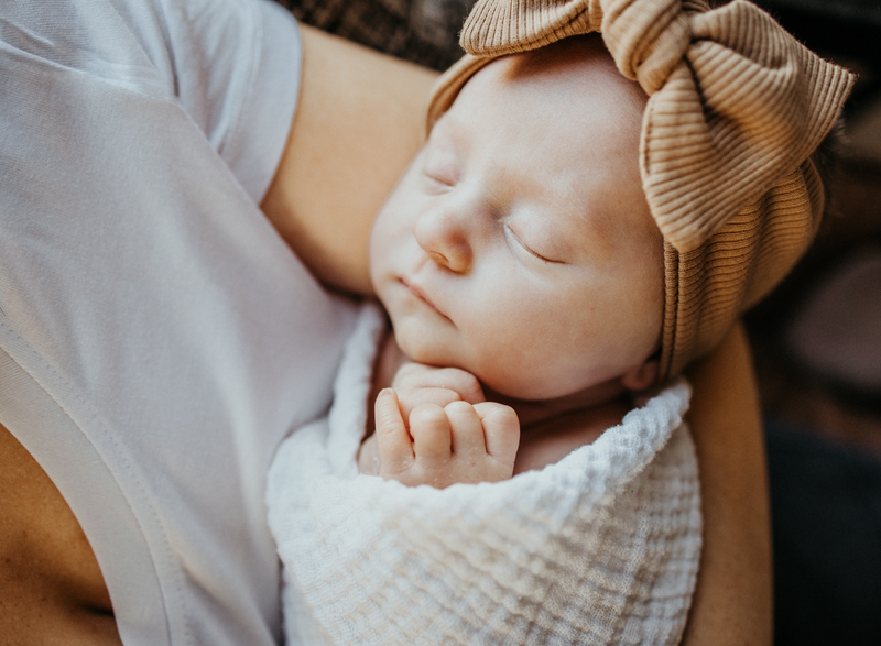 Newborn photographer captures a baby girl sleeping wrapped in a white swaddle and giant brown bow on her head. She is being held in her mother's arms.