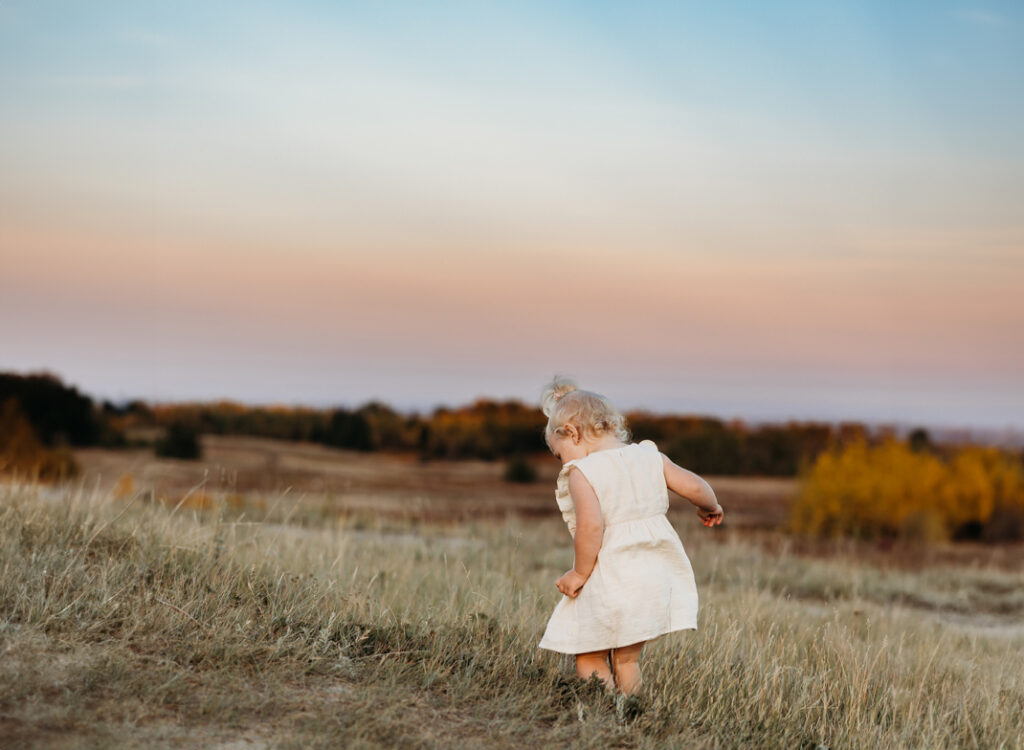 a little girl walks away from the camera in a big field surrounded by trees. The sun set casts a pastel coloured sky with purple, pink, orange, yellow and blue in the background.