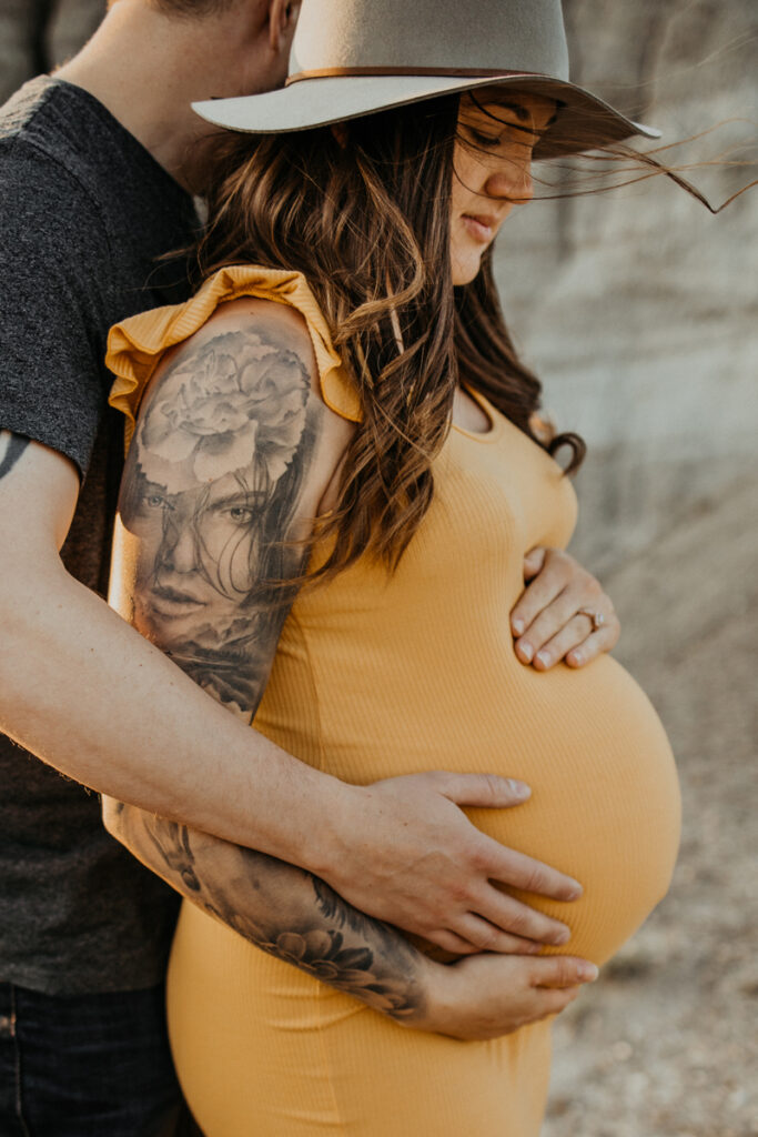 Pregnat woman with long dark hair, a yellow dress, floppy hat and arm sleeve tattoo stand against rocky hills. Dad to be stands behind her holding her baby bump in a maternity photosession